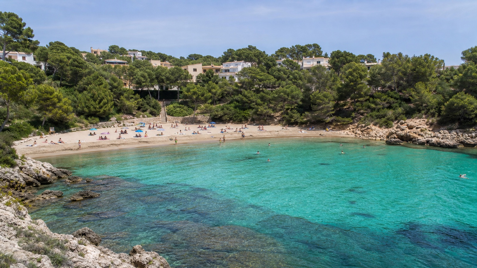 KI-generiertes Stimmungsbild der kompakten Sa Font de sa Cala mit hellem Sand, ruhigem Wasser und niedriger Bebauung